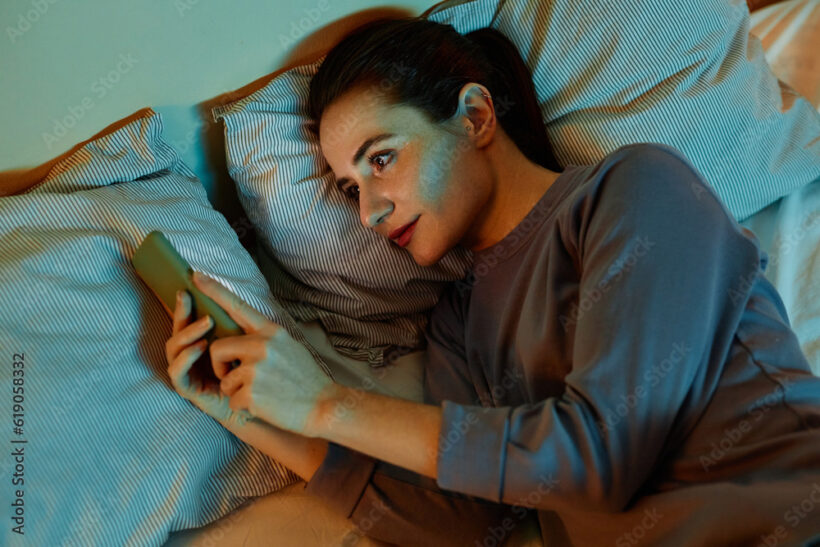 Top view portrait of smiling adult woman using smartphone in bed at night reading text messages and scrolling social media